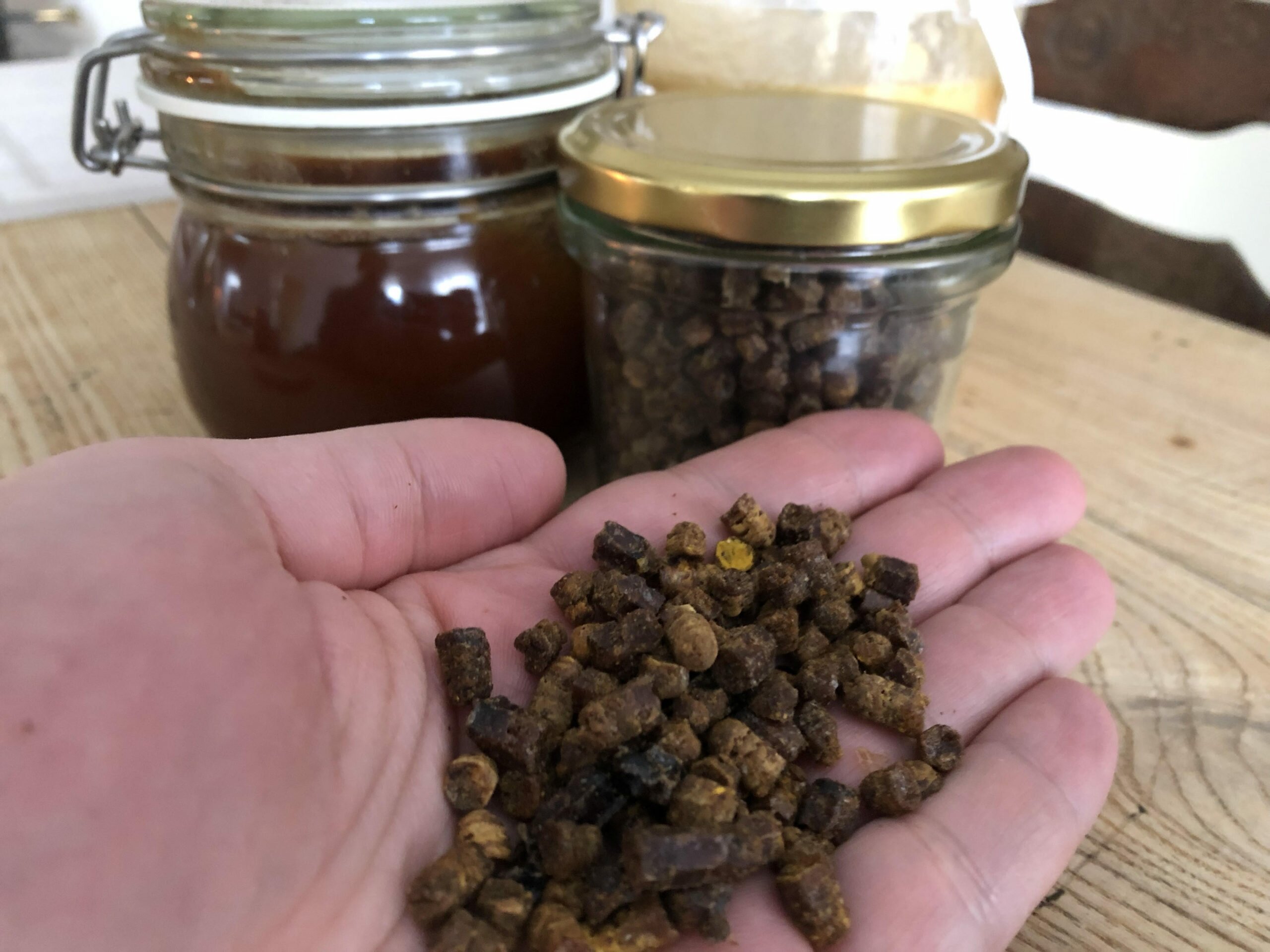 Bee bread in a hand next to three jars with honey and bee bread on a table
